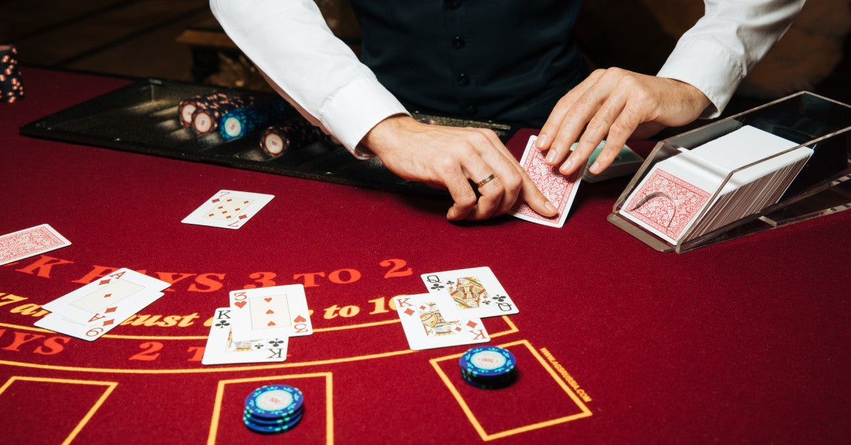 A croupier at a red blackjack casino table deals a card from a card shoe onto a table with multiple players and hands.