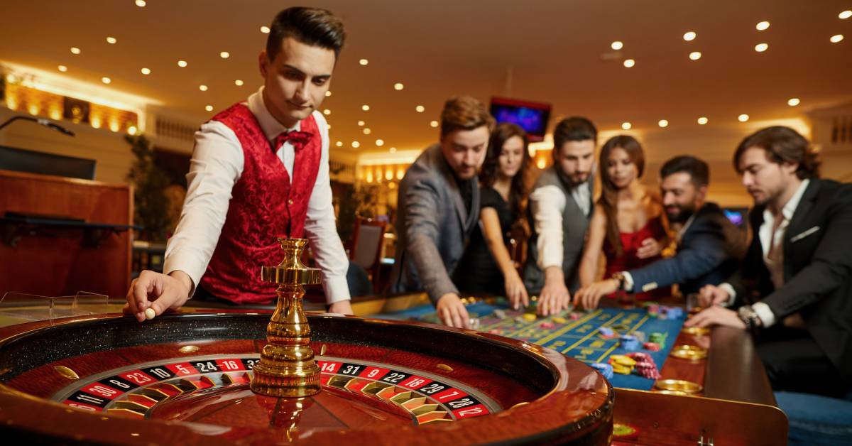 A roulette croupier leans over a roulette wheel to drop the ball while players behind him place their bets.