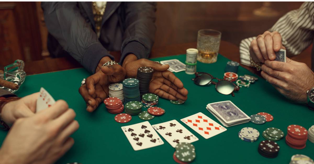 A group of people gathered around a square poker table with stacks of poker chips, cards, and drinks.